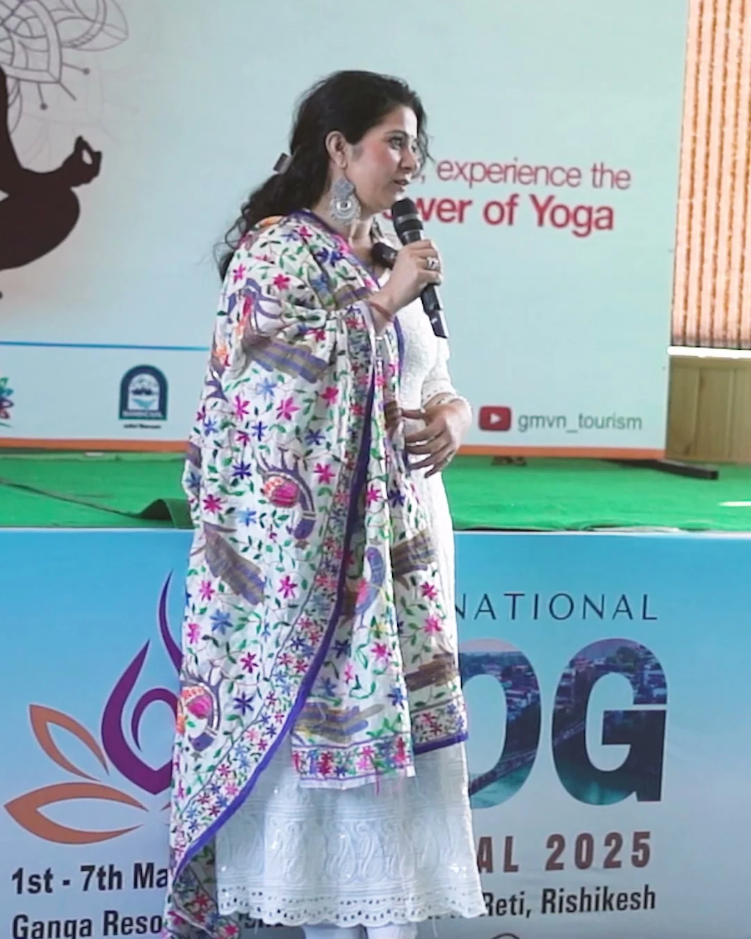 Charu Wadhwa speaking with a microphone on stage at the International Yog Festival 2025 in Rishikesh